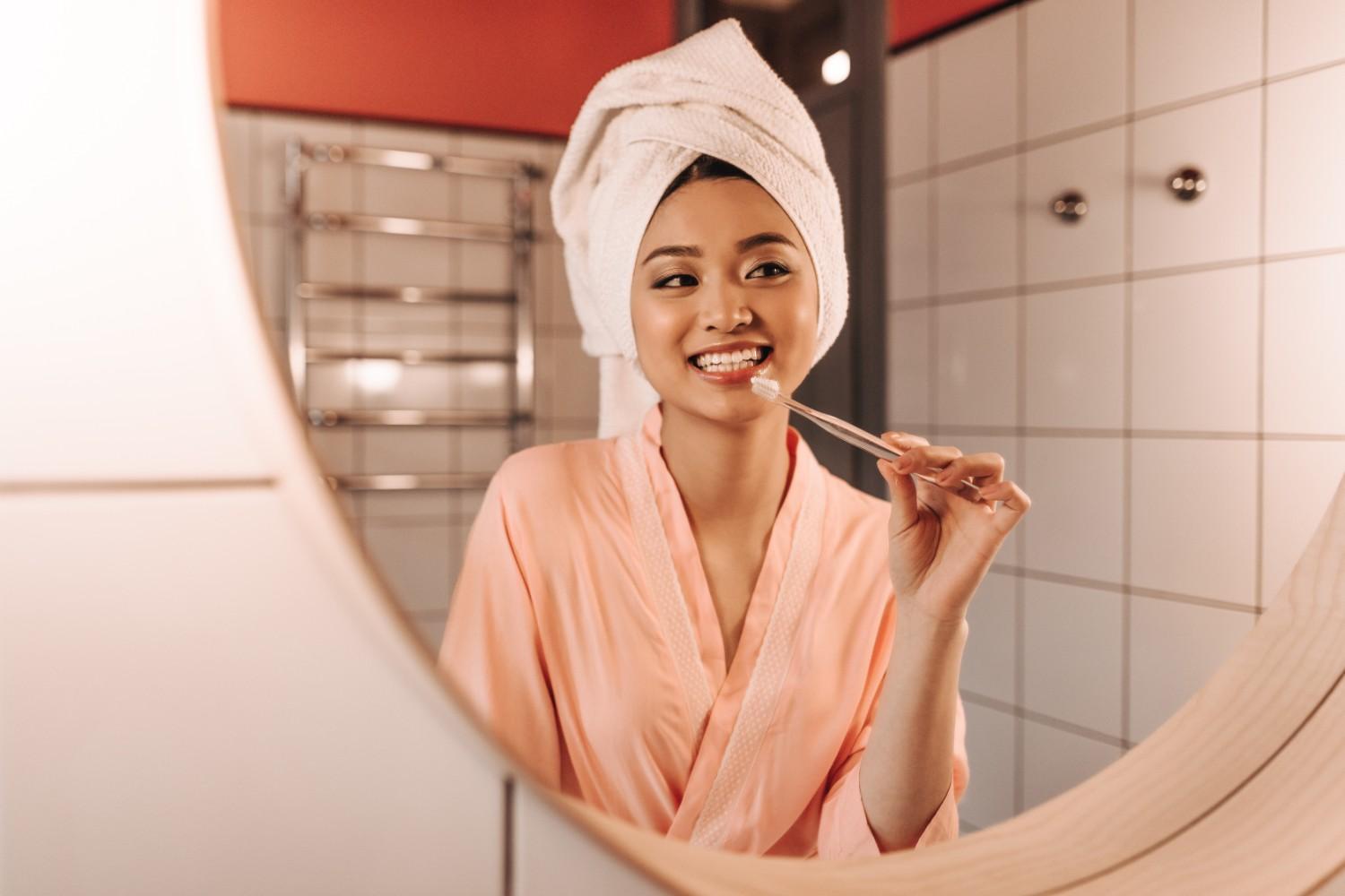 Woman brushing her teeth and smiling in the bathroom mirror.